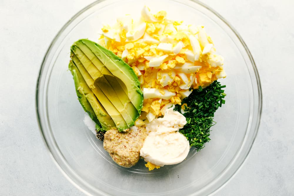 Avocado egg salad is a twist on egg salad adding in the avocado for the color and delicious flavor! Add this to any sandwich or eat it straight from the bowl, it will be a new favorite! The ingredients to make avocado egg salad in a glass mixing bowl.