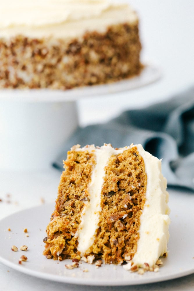 Carrot cake is moist, rich in flavor and melts in your mouth! This scrumptious dessert is what everyone will be raving about! A slice of carrot cake on a plate with the carrot cake in the background on a cake platter.