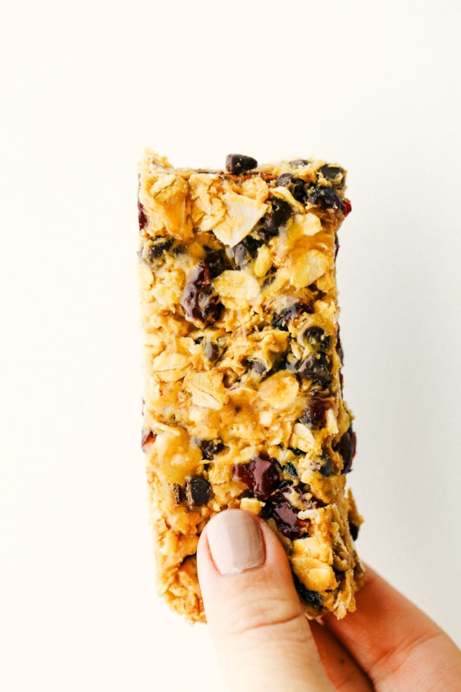 Easy to make chewy granola bars that are homemade filled with regular oats, dried cranberries, salty almonds, mini dark chocolate chips all combined with a sweet honey and a peanut butter flavoring. Snack time just got easier with these tasty bars!ย A granola bar standing up on a photo with a hand holding it.