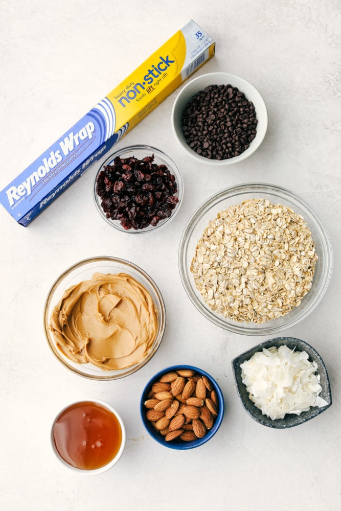 Easy to make chewy granola bars that are homemade filled with regular oats, dried cranberries, salty almonds, mini dark chocolate chips all combined with a sweet honey and a peanut butter flavoring. Snack time just got easier with these tasty bars!ย Granola bar ingredients in small bowls with Reynolds warp non stick aluminum foil in the photo.