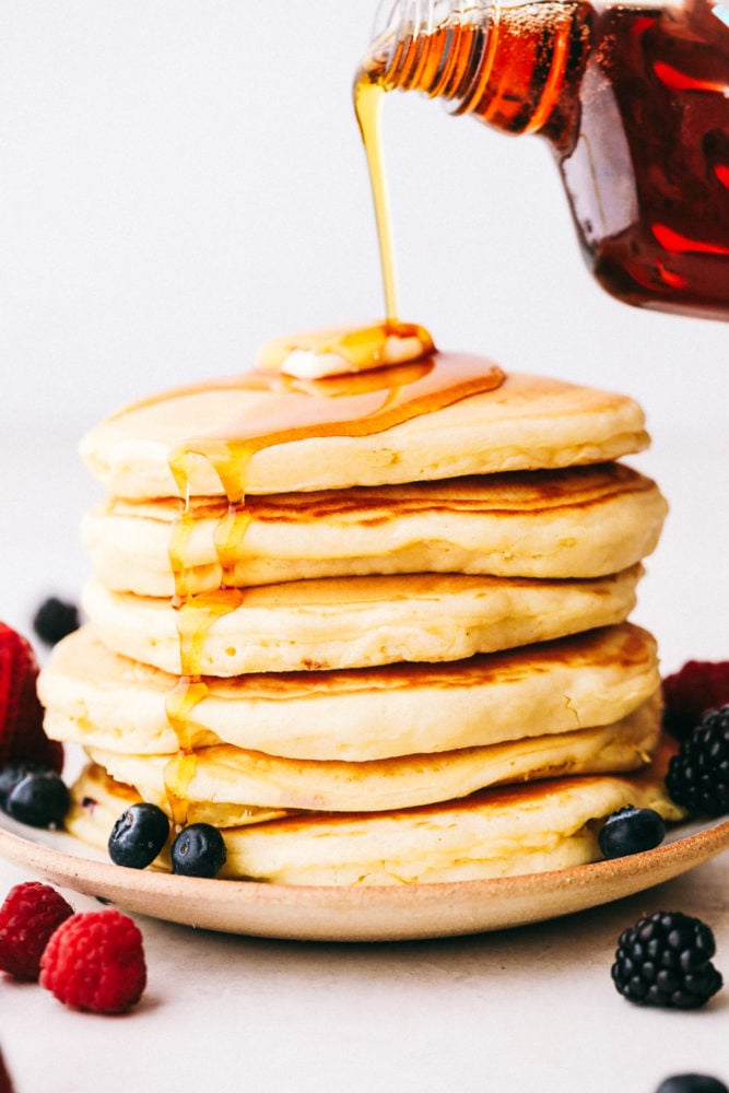Grandma's homemade fluffy pancakes are the ultimate comfort breakfast! They're light and airy, yet satisfyingly filling, making them the perfect morning treat. A stack of 6 pancakes with syrup being poured over top of butter and fresh berries.