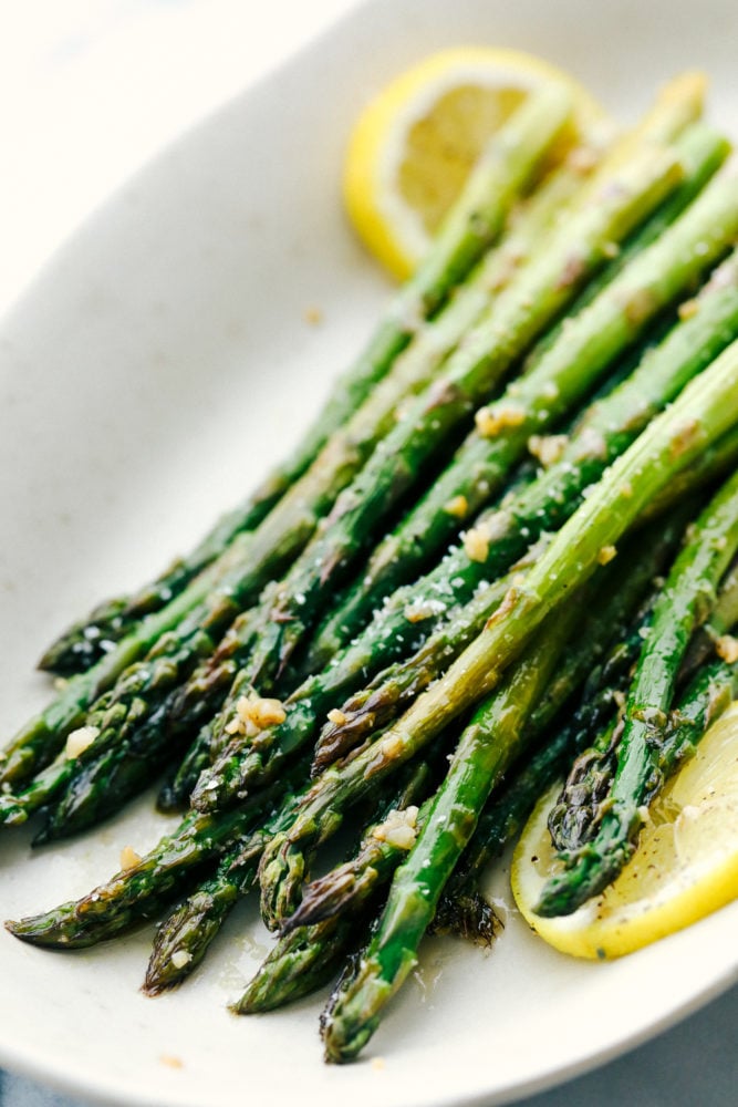 This lemon honey garlic asparagus recipe has a crisp outside with a tender inside cooked in a lemon, honey marinade with minced garlic. This asparagus compliments any main dish and adds a colorful feel to the table! Asparagus placed on a white plate with lemon garnished throughout.