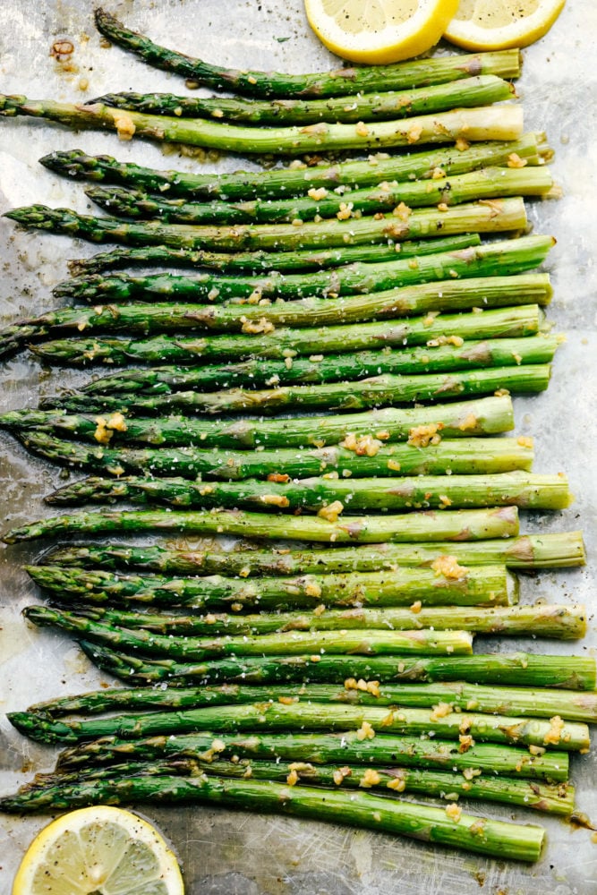 This lemon honey garlic asparagus recipe has a crisp outside with a tender inside cooked in a lemon, honey marinade with minced garlic. This asparagus compliments any main dish and adds a colorful feel to the table! Asparagus all lined up flat on the baking sheet with lemons garnished around them.
