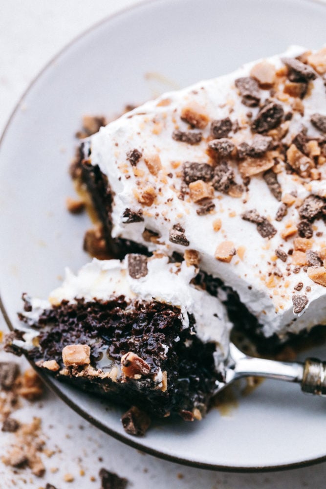 This better than anything cake is a chocolate cake filled with caramel pockets, layered with cool whip and topped with heath bites making ย this cake extremely moist and flavorful in every bite! Make this your next party cake!ย A slice of better than anything cake on a white plate with a fork taking a bite out of the cake.