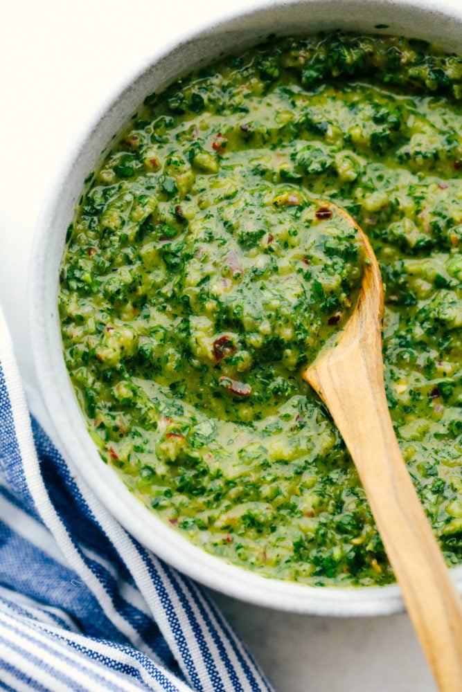 This chimichurri sauce is sweet and savory with a hint of spice and crunch from a tasty red onion. Upgrade your meat or poultry by spreading this flavorful sauce over top creating an intense and delicious flavor every time! Chimichurri sauce in a white bowl with a wooden spoon stirring it.