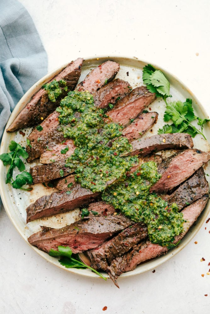 This perfectly grilled flank steak is tender and juicy and topped with the most delicious chimichurri sauce. Combined this main dish is one of our family favorites! Flank steak sliced against the grain with chimichurri sauce on top on a white plate.