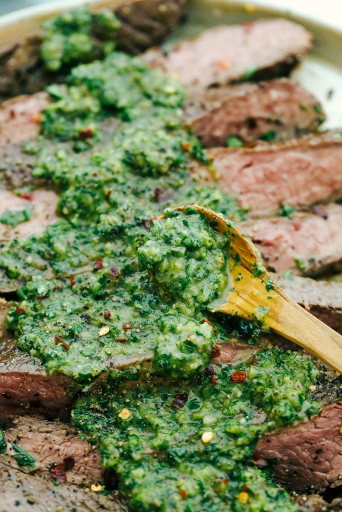 This chimichurri sauce is sweet and savory with a hint of spice and crunch from a tasty red onion. Upgrade your meat or poultry by spreading this flavorful sauce over top creating an intense and delicious flavor every time! Chimichurri sauce laid over steak with a wooden spoon.