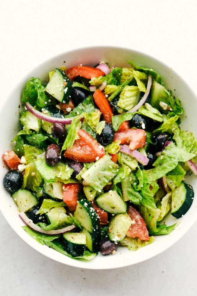 This greek salad dressing is sweet and savory added to any salads, sandwiches or use as a marinade. This dressing is so versatile you will be using it often! Greek Salad Dressing poured over a Greek Salad in a white bowl.