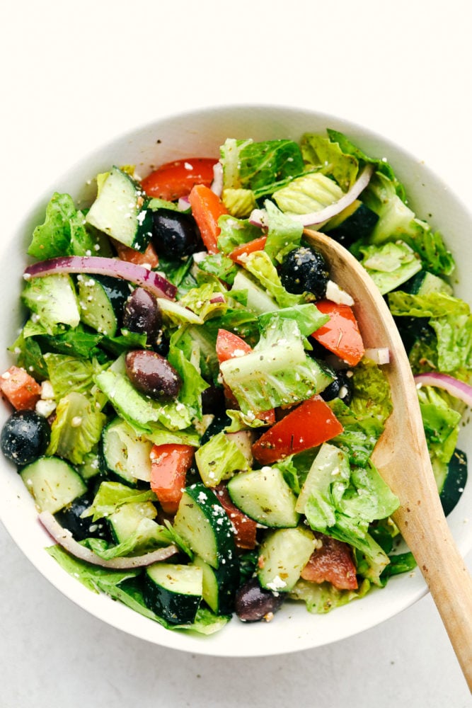 Greek Salad is a fresh, crunchy, vibrant salad with the BEST Greek dressing! Every bite is packed with color and the best flavors that will keep you digging in for more! Greek salad in a white bowl with a wooden spoon.
