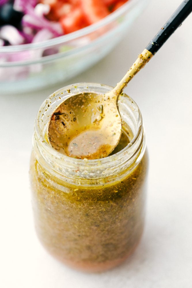 This greek salad dressing is sweet and savory added to any salads, sandwiches or use as a marinade. This dressing is so versatile you will be using it often! Greek Salad Dressing in a jar with a spoon in it.