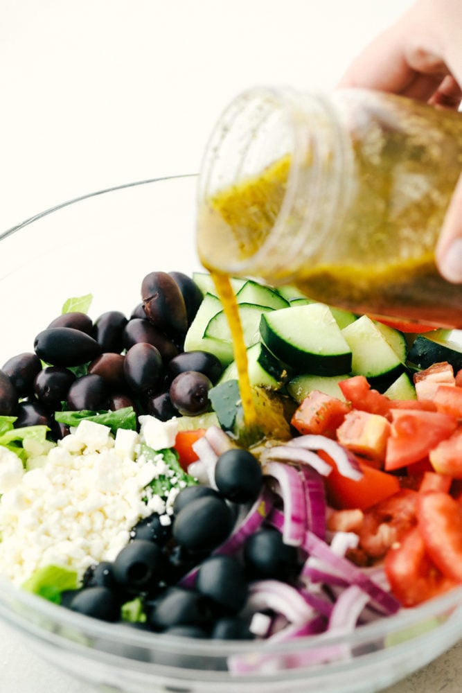This greek salad dressing is sweet and savory added to any salads, sandwiches or use as a marinade. This dressing is so versatile you will be using it often! Greek Salad with olives, feta cheese, tomatoes and cucumbers with Greek Salad Dressing being poured over top.