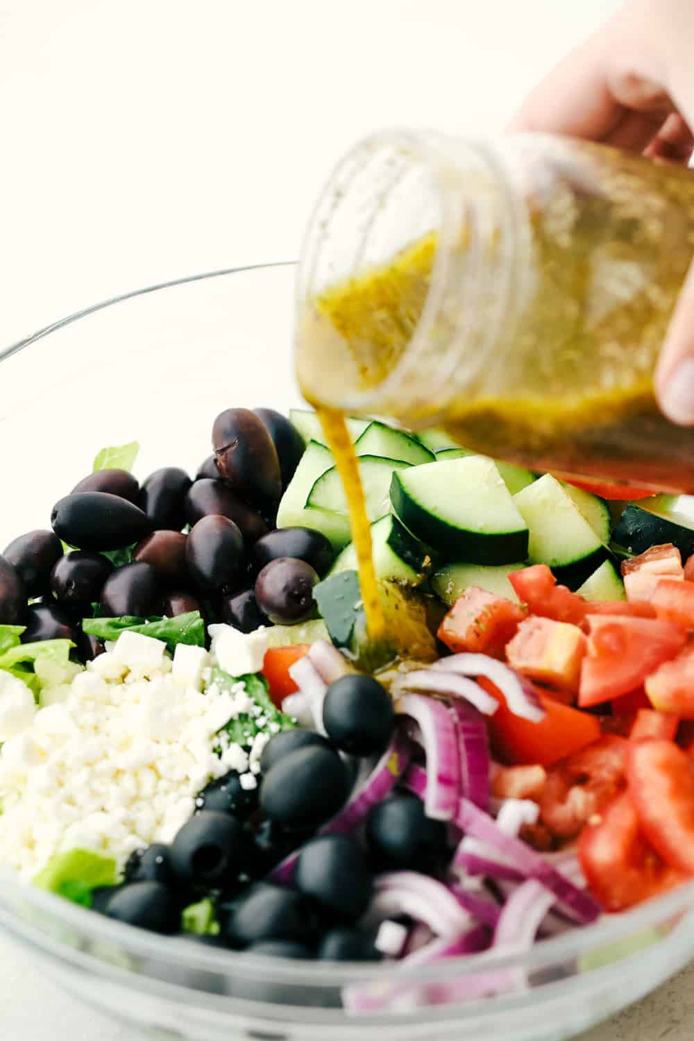 Greek Salad is a fresh, crunchy, vibrant salad with the BEST Greek dressing! Every bite is packed with color and the best flavors that will keep you digging in for more! Pouring dressing on Greek salad ingredients.