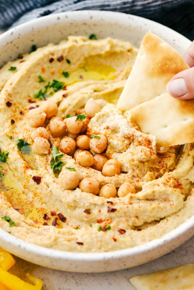 This is the BEST hummus that is fresh, flavorful, smooth and creamy plus quick and easy to make. Add this hummus for your next dip! Hummus in a white bowl with a pita chip being dipped in it and chickpeas over top as a garnish.