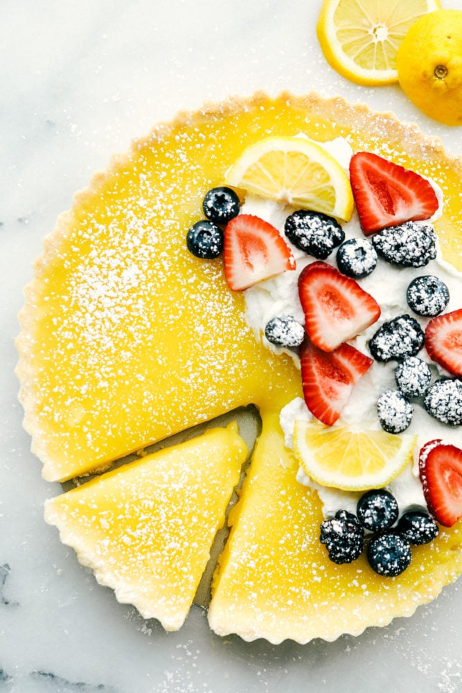 This sweet and savory lemon tart dessert is pure heaven and melts in your mouth with every bite! From the buttery shortbread crust, to the sweet and creamy lemon curd then topped with fresh fruit and powdered sugar, this dessert is rich and flavorful. Lemon tart photo with fresh berries on top with lemon slices and powder sugar sprinkled on top and a slice cut out of the tart.