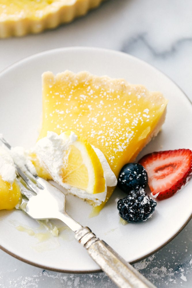 This sweet and savory lemon tart dessert is pure heaven and melts in your mouth with every bite! From the buttery shortbread crust, to the sweet and creamy lemon curd then topped with fresh fruit and powdered sugar, this dessert is rich and flavorful. Lemon tart on a white plate with fresh berries on the side and a bite taken out of the tart.