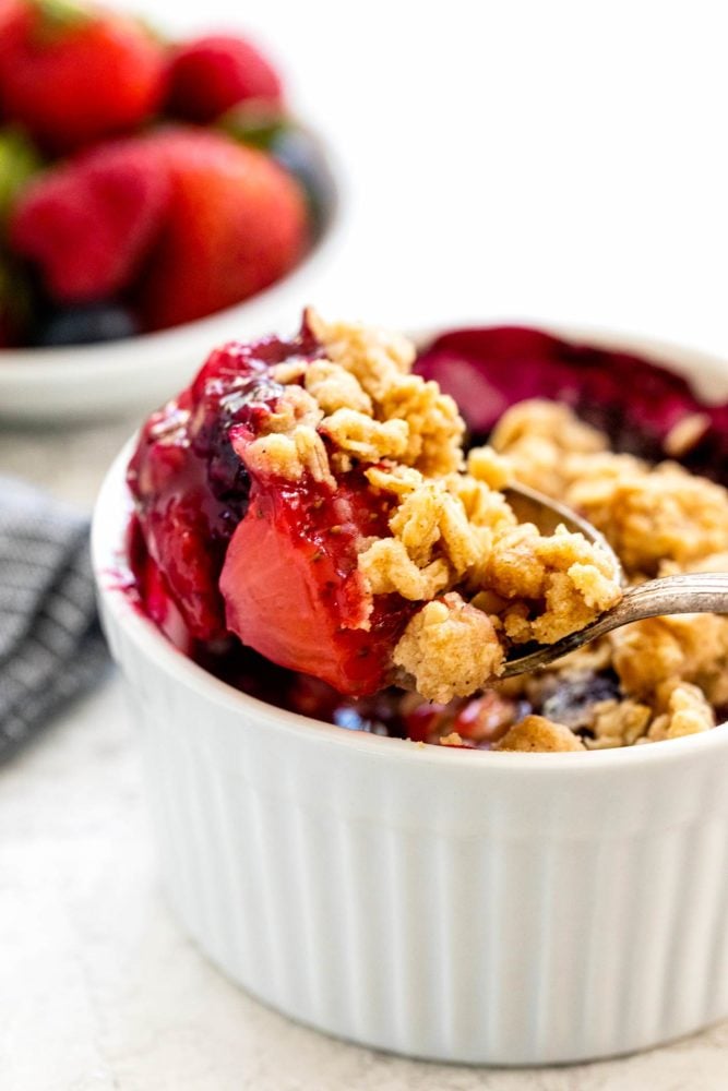 Mixed berry crisp is a colorful combination of fruit with a crunchy oat topping. These smaller individual portions baked in a ramekin make it easy to serve. The warm dessert tastes incredible topped with whipped cream or ice cream. spoon scooping into a dessert
