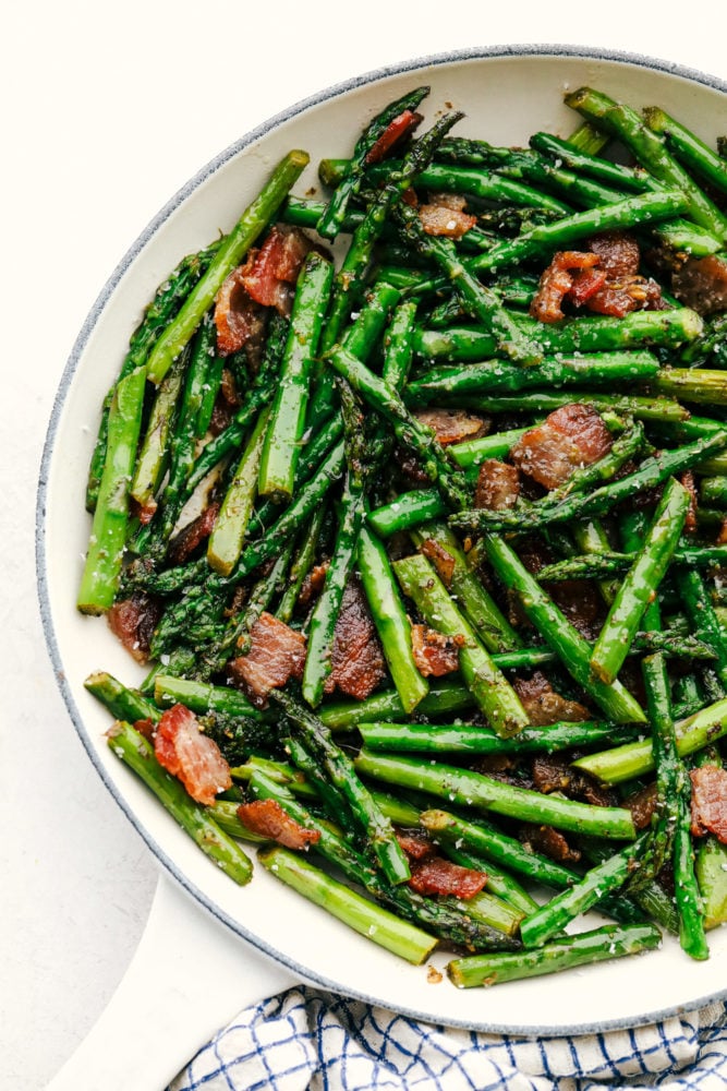 This garlic asparagus is an easy, fresh and tender side dish with bacon chopped up and sautéed together. Adding bacon enhances this delicious dish to the next level! Garlic asparagus with bacon in a white skillet.