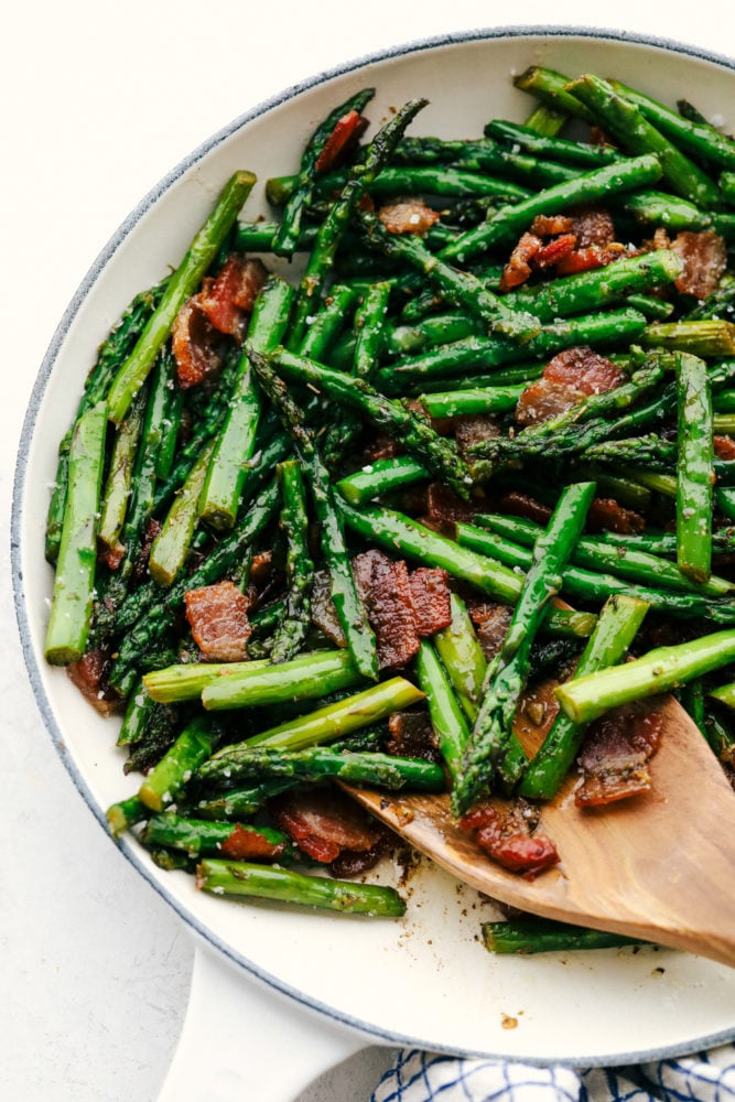 This garlic asparagus is an easy, fresh and tender side dish with bacon chopped up and sautéed together. Adding bacon enhances this delicious dish to the next level! Garlic asparagus with bacon being stirred in a skillet with a wooden spoon.