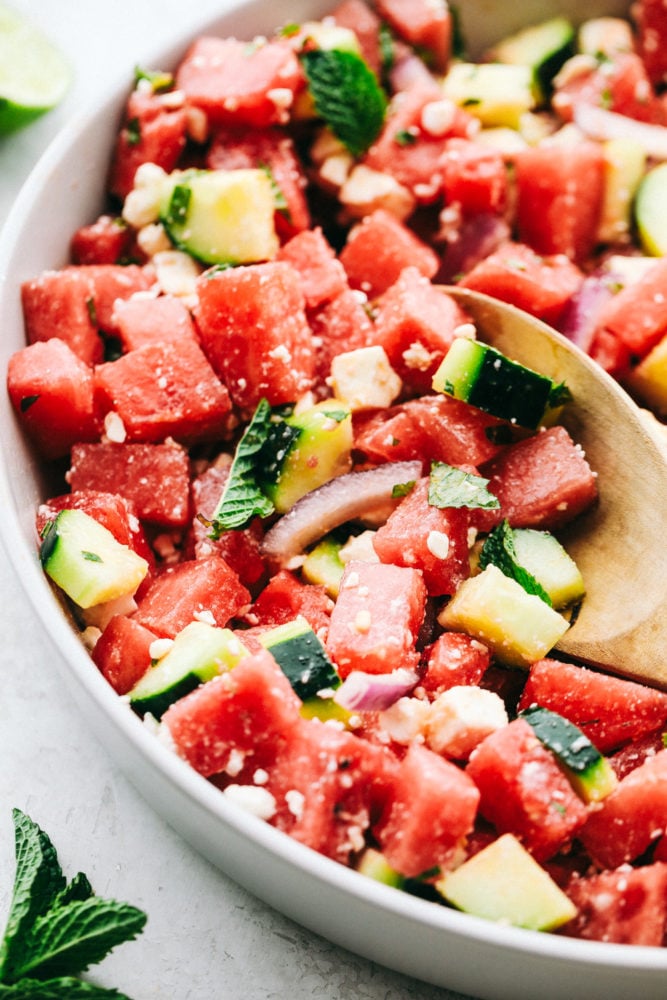 Watermelon Salad is bursting with flavors from the juicy watermelon and salty feta cheese, then topped off with hints of lime and mint.ย It is the perfect fresh sweet and savory mix of flavors that just scream summer!! Watermelon salad in a white bowl with a wooden spoon in the salad.