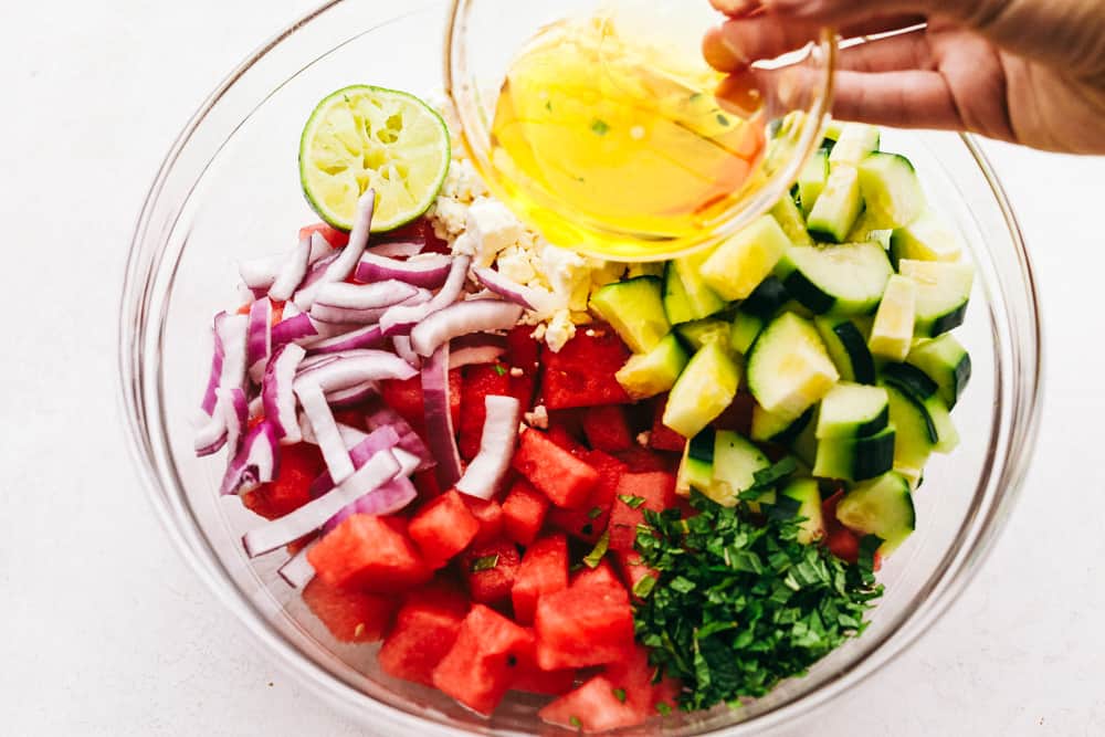 Watermelon Salad is bursting with flavors from the juicy watermelon and salty feta cheese, then topped off with hints of lime and mint.ย It is the perfect fresh sweet and savory mix of flavors that just scream summer!! Ingredients of the salad in a glass bowl separated and having the dressing being poured overtop.