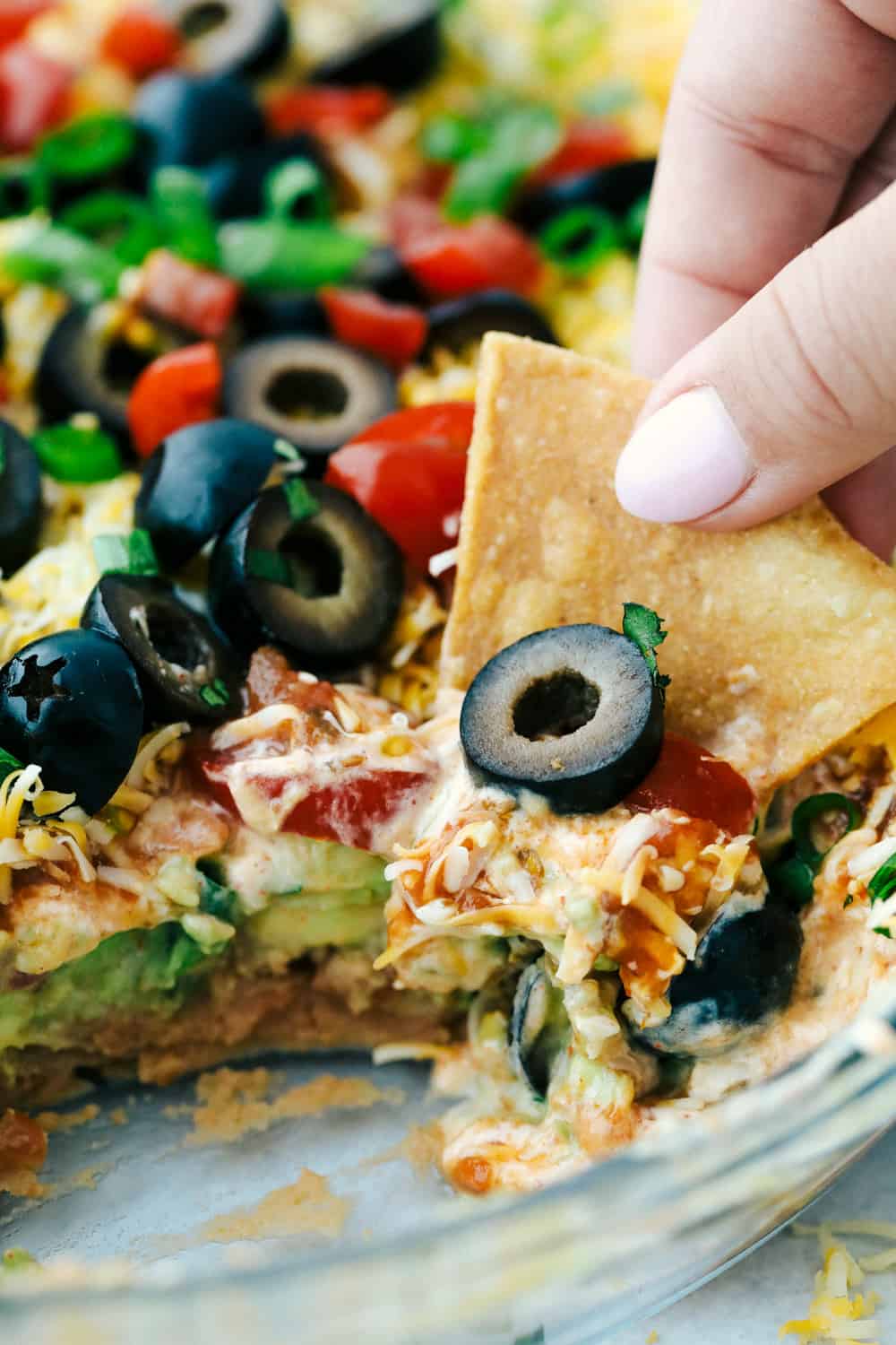 Awesome 7 Layer Dip is an addicting creamy dip that includes all your favorites, like guacamole, refried beans, and sour cream! You will be amazed at how simple it is to put together such an incredibly delicious dip! Close up shot of someone using a tortilla chip to scoop out a bite,
