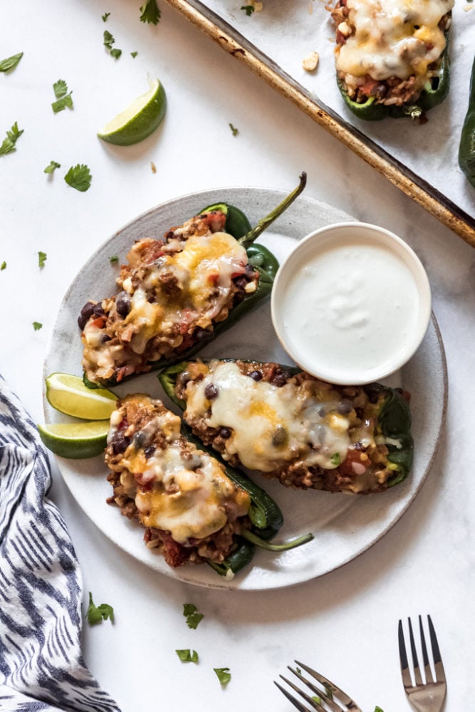 Stuffed Poblano Peppers are loaded with southwest seasoned ground beef, rice, veggies, and cheese for a satisfying dinner with just a little bit of a kick! Everybody raves about this twist on classic stuffed peppers! Three stuffed poblano peppers on a plate with a bowl of crema and lime wedges.
