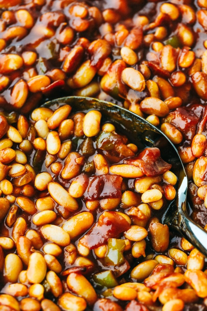 ย I am showing you how to make the best baked beans from start to finish. ย Making homemade baked beans is a lot easier than you think! Close up on finished baked beans being scooped with a serving spoon.