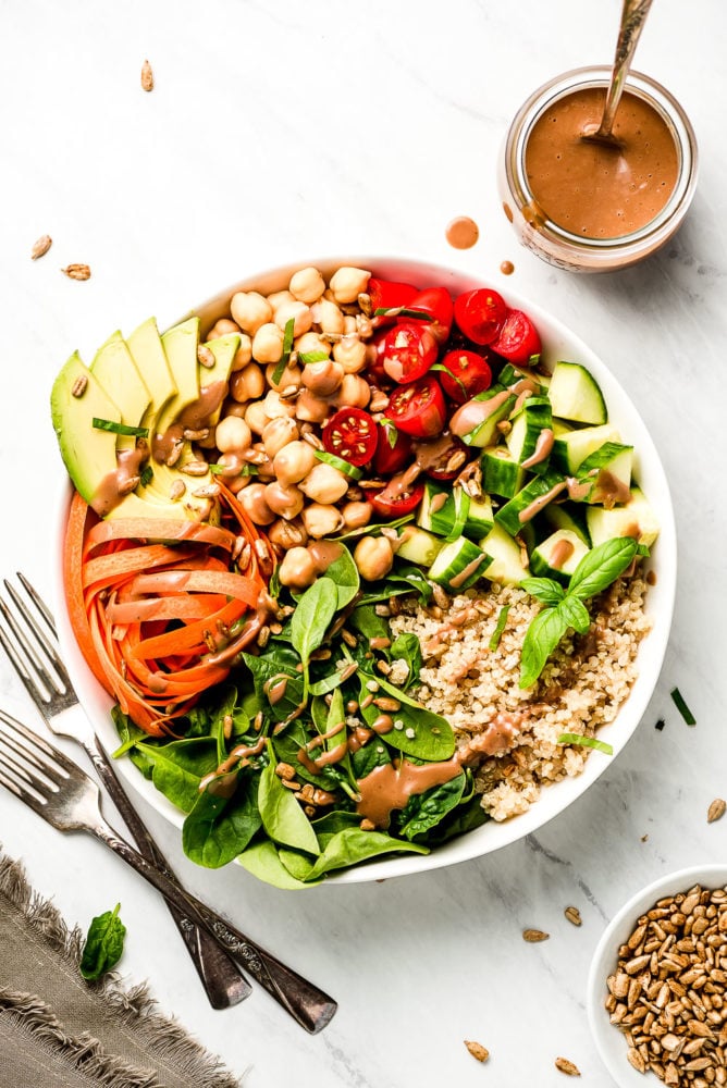 This Vegan Buddha Bowl is loaded with colorful raw summer vegetables, whole grains, good fats, and topped with a tangy creamy balsamic dressing. Make adjustments to use up whatever vegetables you have on hand and enjoy this delicious vegan meal for lunch or dinner all summer long! A raw vegetable Vegan Buddha Bowl made up of with spinach, carrots, avocado, chickpeas, grape tomatoes, cucumbers, quinoa, basil, sesame seeds, and a creamy balsamic dressing.