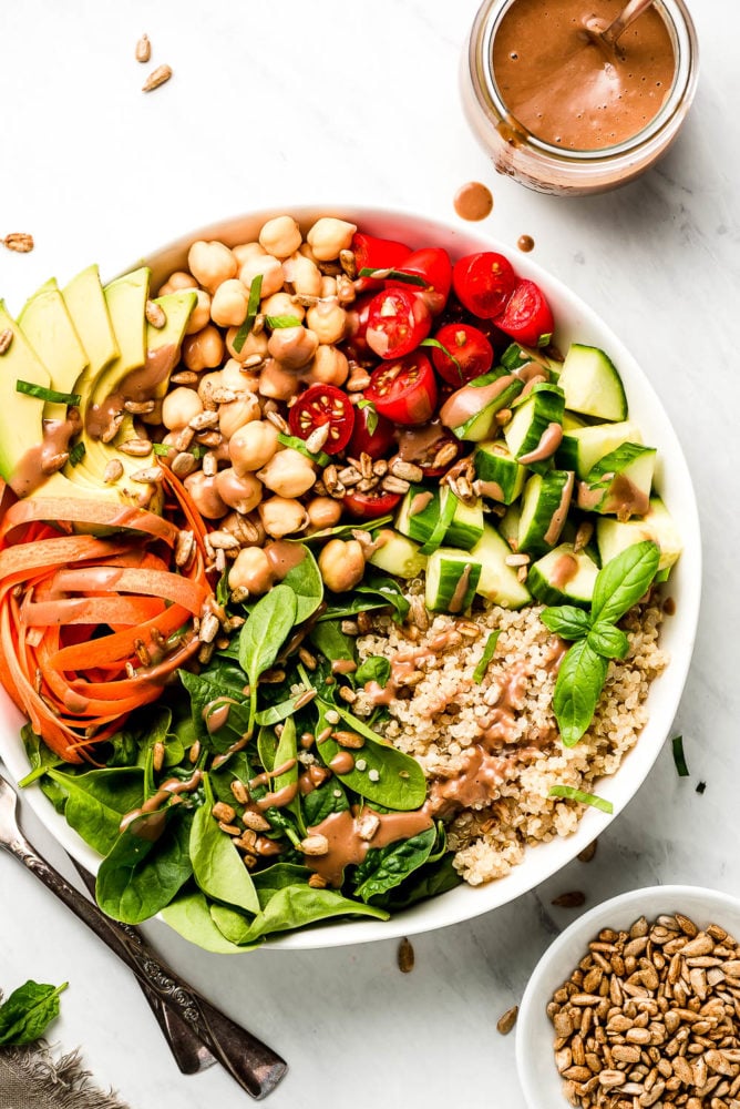 This Vegan Buddha Bowl is loaded with colorful raw summer vegetables, whole grains, good fats, and topped with a tangy creamy balsamic dressing. Make adjustments to use up whatever vegetables you have on hand and enjoy this delicious vegan meal for lunch or dinner all summer long! A large Buddha Bowl with spinach, quinoa, carrots, cucumbers, tomatoes, chickpeas, and avocado with a jar or dark creamy dressing to the side.