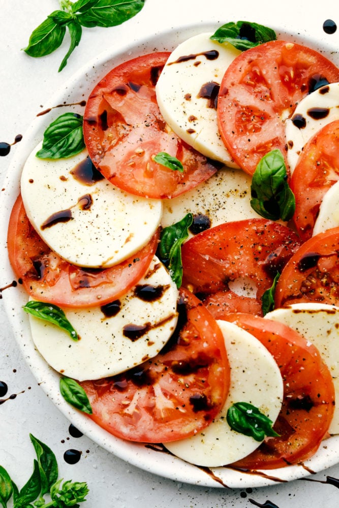 Caprese Salad is made with layers of fresh tomato slices, basil, and mozzarella cheese, then drizzled with balsamic glaze and topped with a sprinkle of salt and pepper. It is the perfect summer meal that you will crave all year! Caprese Salad on a white plate.