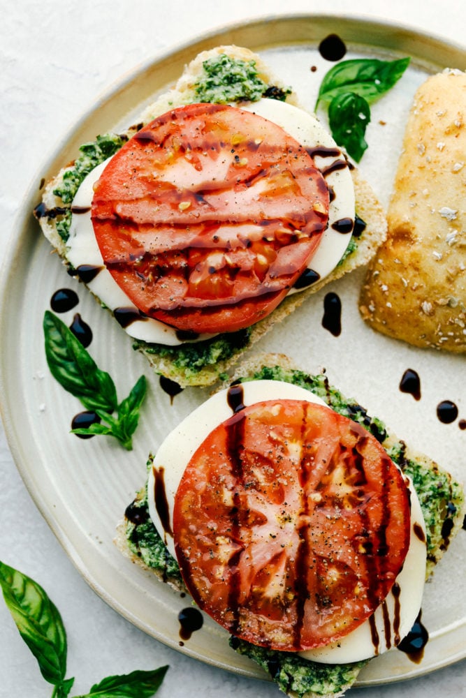 A Caprese Sandwich with Basil Pesto is a thick ciabatta bread stuffed with layers of tomato, mozzarella, and fresh pesto.ย Add a drizzle of a balsamic glaze and you will have a homemade sandwich that is better than anything you could buy at a restaurant! Balsamic glaze drizzled over caprese sandwiches on a plate.