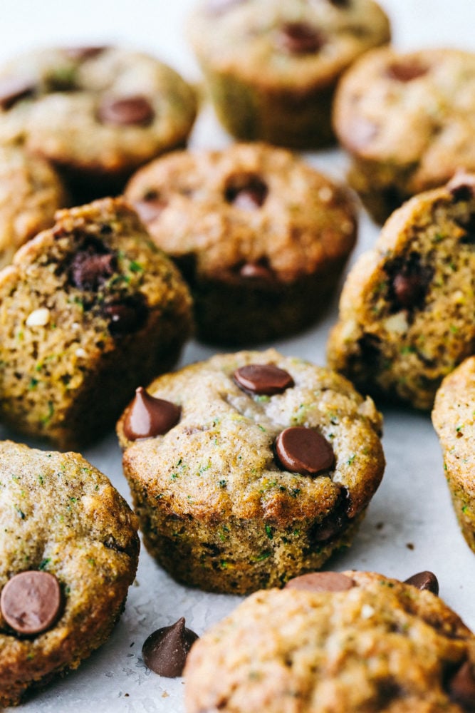 Chocolate Chip Zucchini Muffins are moist and rich muffins that are loaded with flavor from rich chocolate and shredded zucchini!ย They will be a huge hit and will give you a great way to use all that zucchini from your garden! Scattered chocolate chip zucchini muffins sitting on parchment paper.