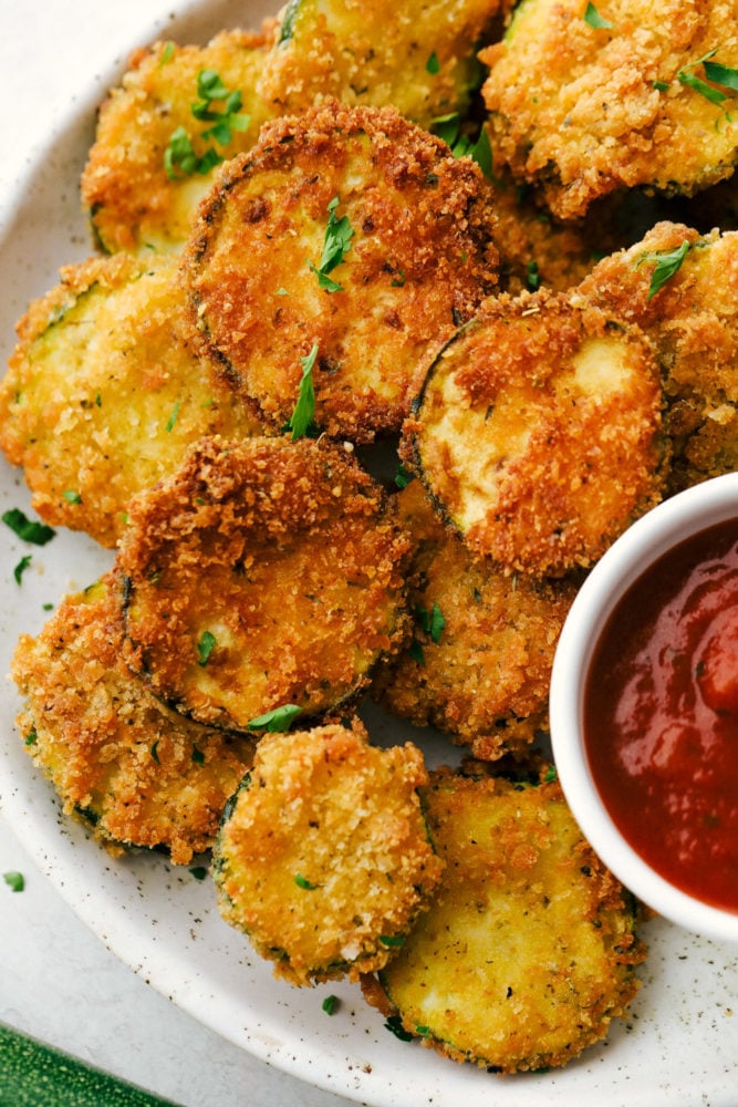 Crispy Parmesan Fried Zucchini is made with savory parmesan and panko coated zucchini bites. They will quickly become a favorite for an easy and crowd-pleasing snack, appetizer or side dish! Crispy Parmesan Fried Zucchini on a white plate with marinara sauce.