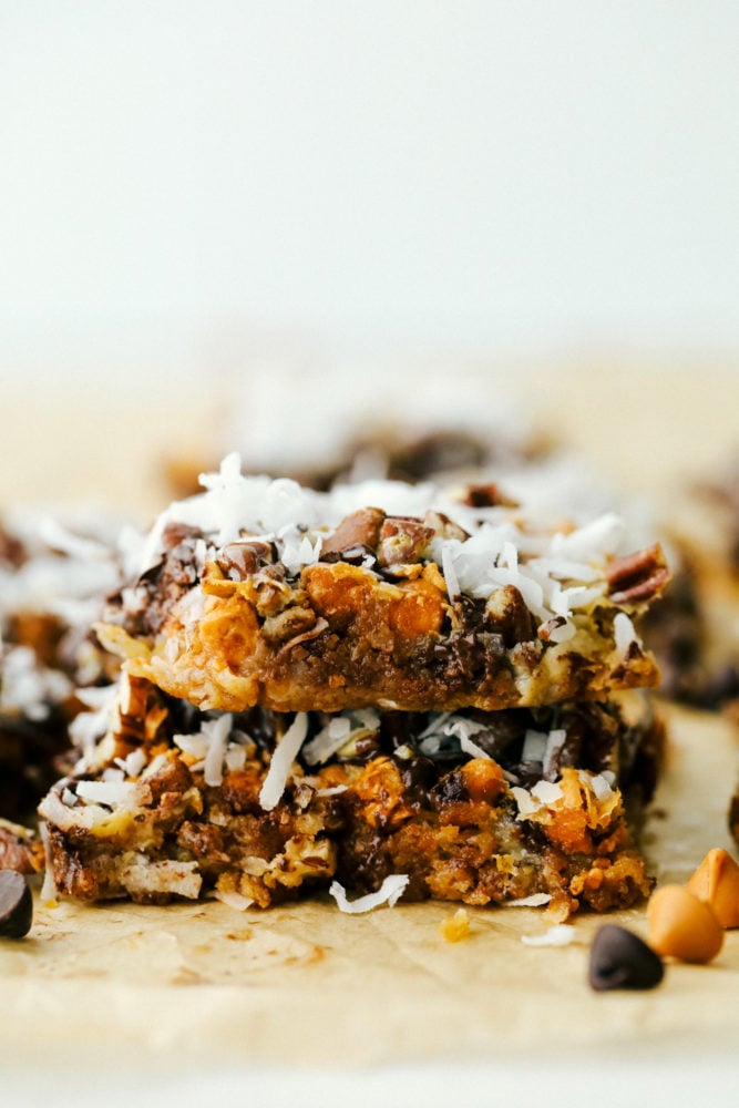 7 Layer Magic Bars are loaded with layers of rich flavors from coconut, pecans, butterscotch, and chocolate. You will be amazed how easy it is to create a bar this delicious where every single bite is ooey gooey perfection! Close up on a stack of 2 seven layer bars.