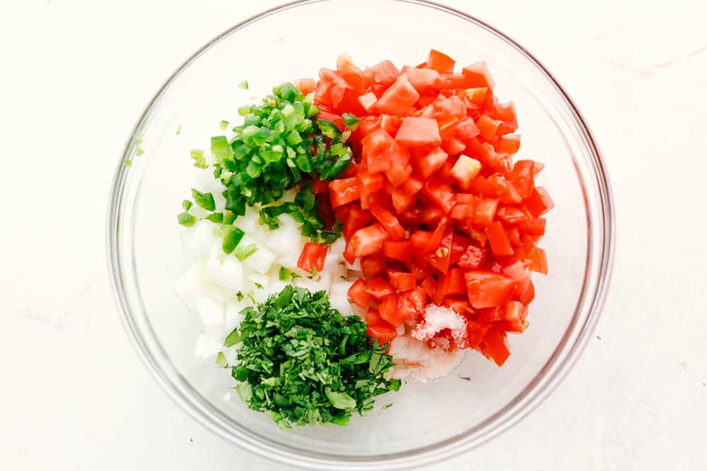 Pico de Gallo is a made from scratch salsa that is loaded with bold flavors from jalapeno, lime, cilantro, and juicy chunks of tomato and onion. One dip will certainly not be enough and you will be scraping the bottom of the bowl in no time! Ingredients for pico de gallo in a clear bowl.