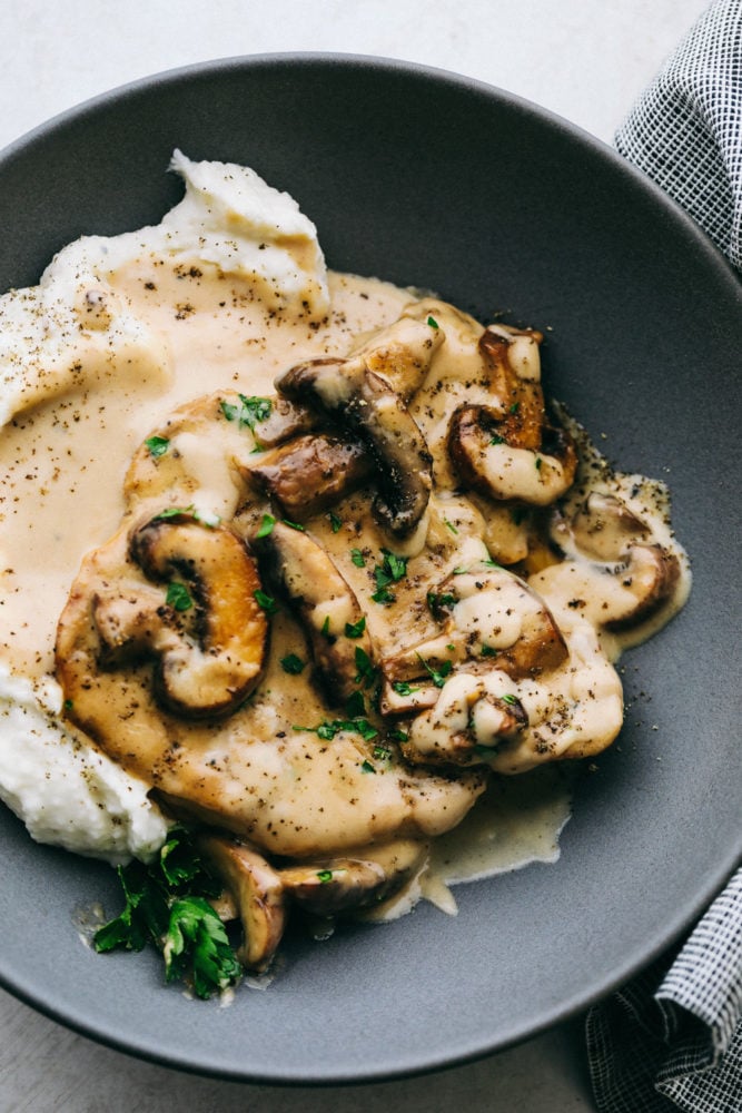 Smothered Pork Chops in an Amazing Mushroom Gravy are tender, savory, and satisfying.ย This one pan dinner is the perfect comforting dinner at home that your family will love! Smothered pork chops plated with mashed potatoes on a black plate.