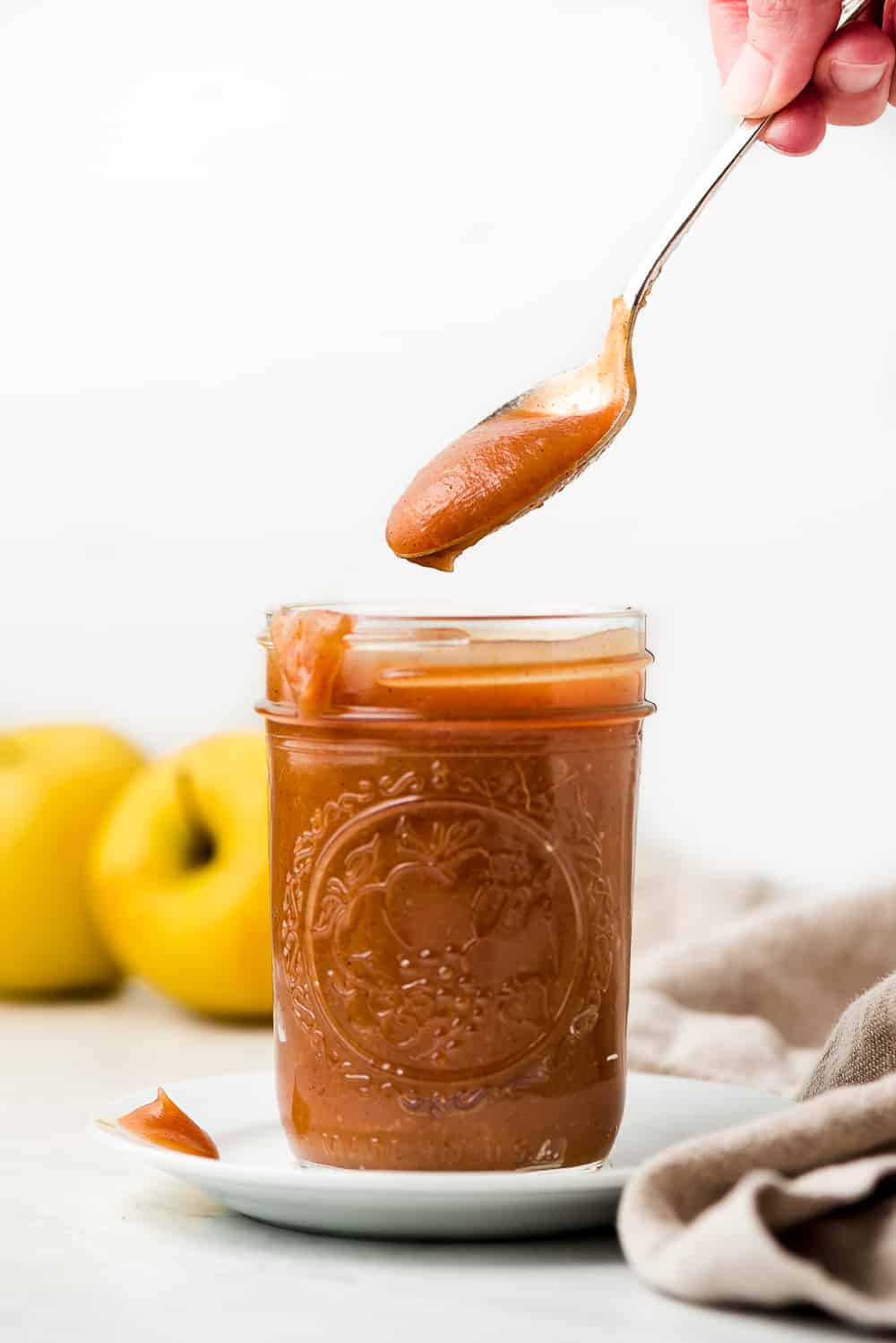 Apple Butter is a sweet velvety concentrated apple spread that youโll want to put on everything. It requires just a few ingredients and a little bit of patience to make this yummy jam like condiment. Lifting a spoonful of Apple Butter out of a jar on a plate with apples in the background.