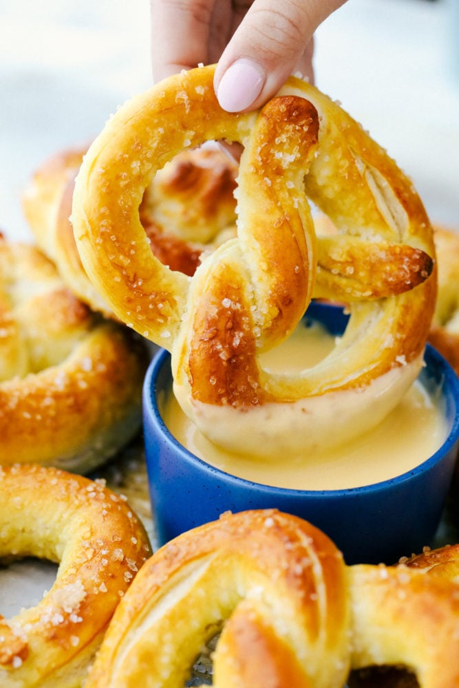 Baked Soft Pretzels are soft and chewy on the inside with the perfect salty golden exterior.ย This recipe will walk you through these easy homemade pretzels with step by step instructions! Dipping a pretzel in cheese sauce.