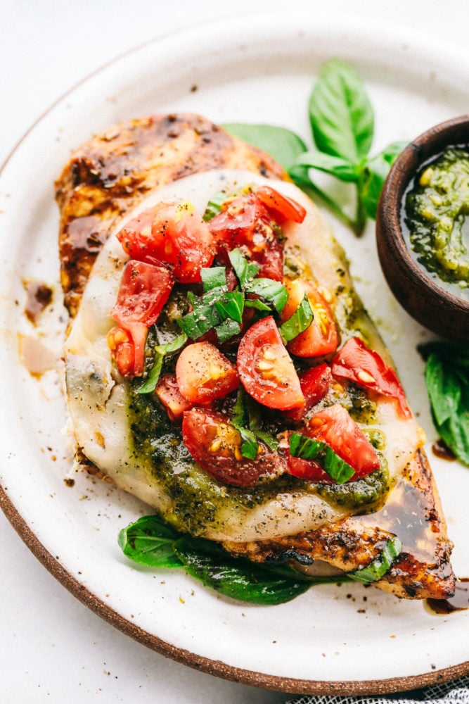 Chicken Margherita is tender and flavor packed grilled chicken that is smothered with mozzarella, pesto, and juicy cherry tomatoes. Liven up dinnertime with this restaurant quality meal that's quick and easy to make at home! Chicken Margherita on a white plate.