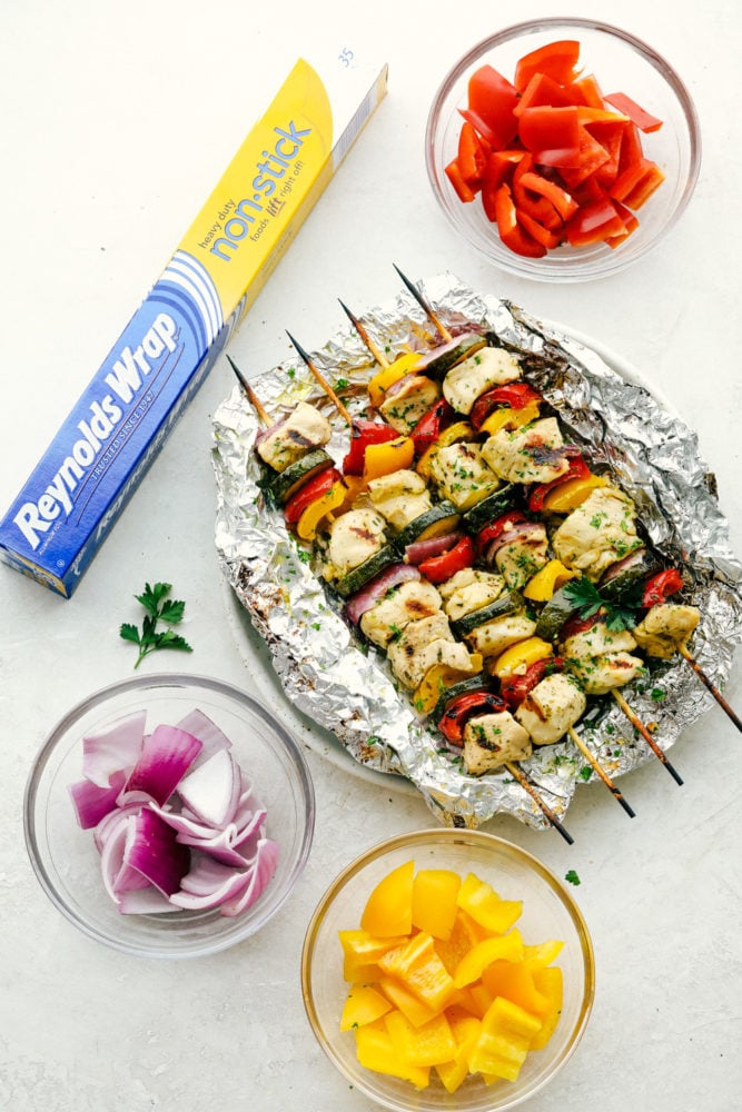 Garlic Ranch Chicken Skewers are the perfect way to enjoy kabobs this summer. Get grilling with this easy to make chicken that has all the flavor on each skewer! Chicken skewers laying on Reynolds wrap with bell peppers and onions in separate glass bowls.