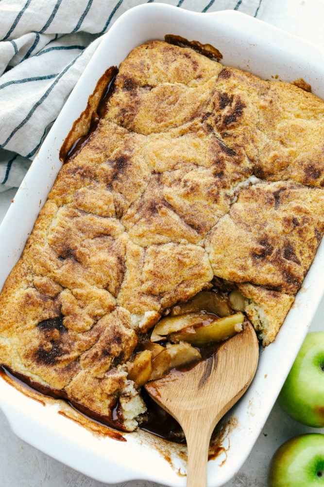 Old Fashioned Apple Cobbler is a warm baked fruit dessert that is topped with a perfect biscuit-like crust.ย The warm cinnamon spice is comforting and satisfying and nails a perfect summer dessert! Scooping apple cobbler out of a white dish.