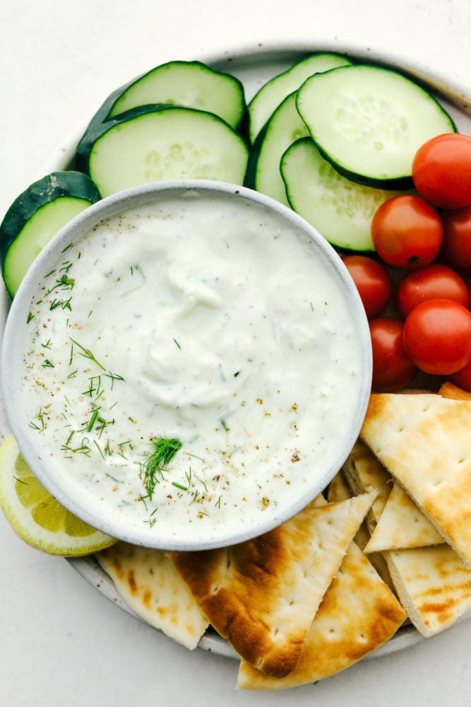 Tzatziki Sauce is a Greek inspired, yogurt-based sauce you and your family will go crazy over. It is creamy and bursting with light and refreshing cucumber, dill, and garlic flavors that will make this a Mediterranean inspired favorite this summer! Close up on tzatziki sauce in a bowl on a veggie platter with pita.