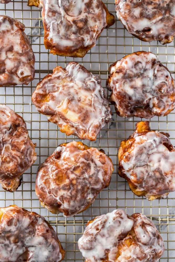 Juicy chunks of apple, coated in batter and shallow fried, these homemade apple fritters are a tender, delicious apple breakfast treat that are also incredibly simple to make. They’re topped off with an addictive but simple icing which takes them to a whole ‘nother level. Top down view of apple fritters on a gold wire rack