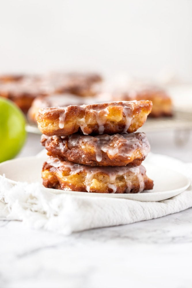 Juicy chunks of apple, coated in batter and shallow fried, these homemade apple fritters are a tender, delicious apple breakfast treat that are also incredibly simple to make. They’re topped off with an addictive but simple icing which takes them to a whole ‘nother level. A stack of 3 apple fritters on a white plate with more in the background