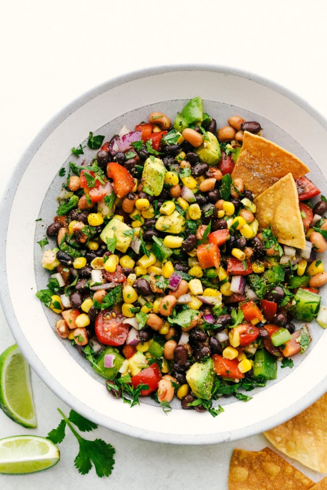 Cowboy caviar is a delicious blend of canned beans, corn and fresh Roma tomatoes, onions and avocados all chopped and prepared to be mixed together to start dipping in. This appetizer is easy to make and has a fresh and flavorful blend! Cowboy caviar in a white bowl with two chips sticking out of the dip.
