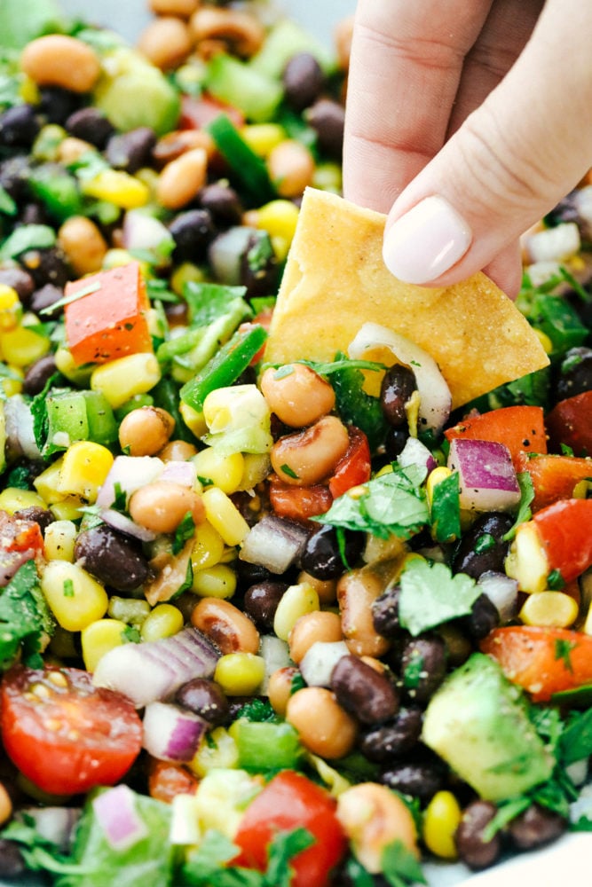 Cowboy caviar is a delicious blend of canned beans, corn and fresh Roma tomatoes, onions and avocados all chopped and prepared to be mixed together to start dipping in. This appetizer is easy to make and has a fresh and flavorful blend! An unclose photo of the caviar and a person dipping in the salsa with a thick, crunchy chip.