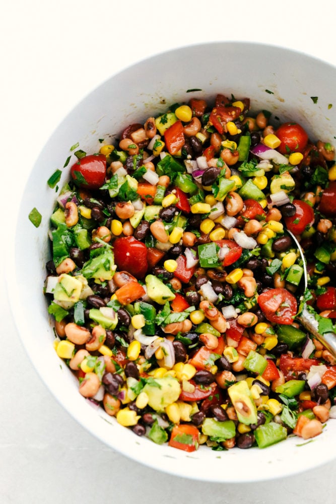 Cowboy caviar is a delicious blend of canned beans, corn and fresh Roma tomatoes, onions and avocados all chopped and prepared to be mixed together to start dipping in. This appetizer is easy to make and has a fresh and flavorful blend! Cowboy caviar in a white bowl.