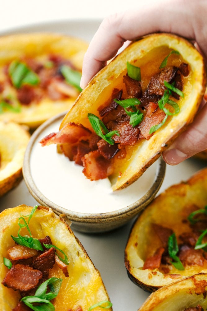Baked Potato Skins have a crispy outer shell that is loaded with a cheddar cheese, bacon and green onion combination. These potatoes are everyones favorite appetizer or side dish! A potato skin being dipped in ranch sauce.