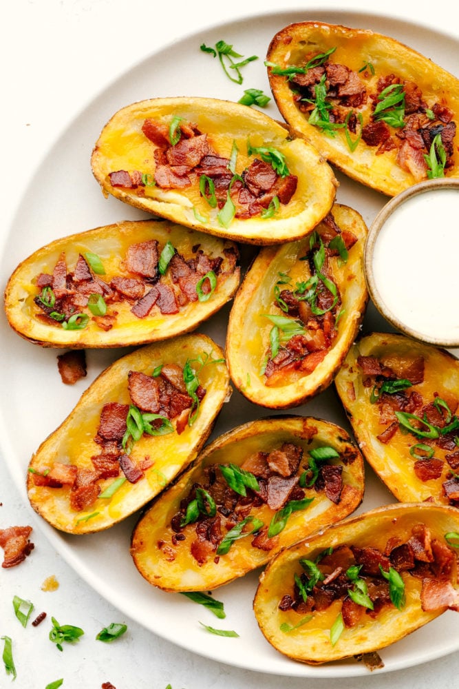 Baked Potato Skins have a crispy outer shell that is loaded with a cheddar cheese, bacon and green onion combination. These potatoes are everyones favorite appetizer or side dish! Potato skins on a white plate with ranch dipping sauce on the side.