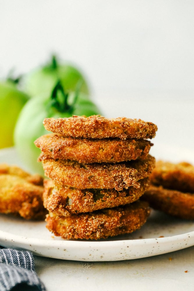 Fried Green Tomatoes are extremely popular sliced and fried crunchy bites of tangy green tomatoes. They are totally addictive and easy to make at home! Stack of fried green tomatoes on a white plate.
